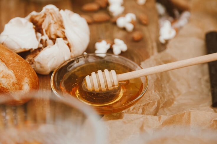 Close-up of a honey dipper in a bowl surrounded by garlic, bread, almonds, and popcorn on parchment paper.