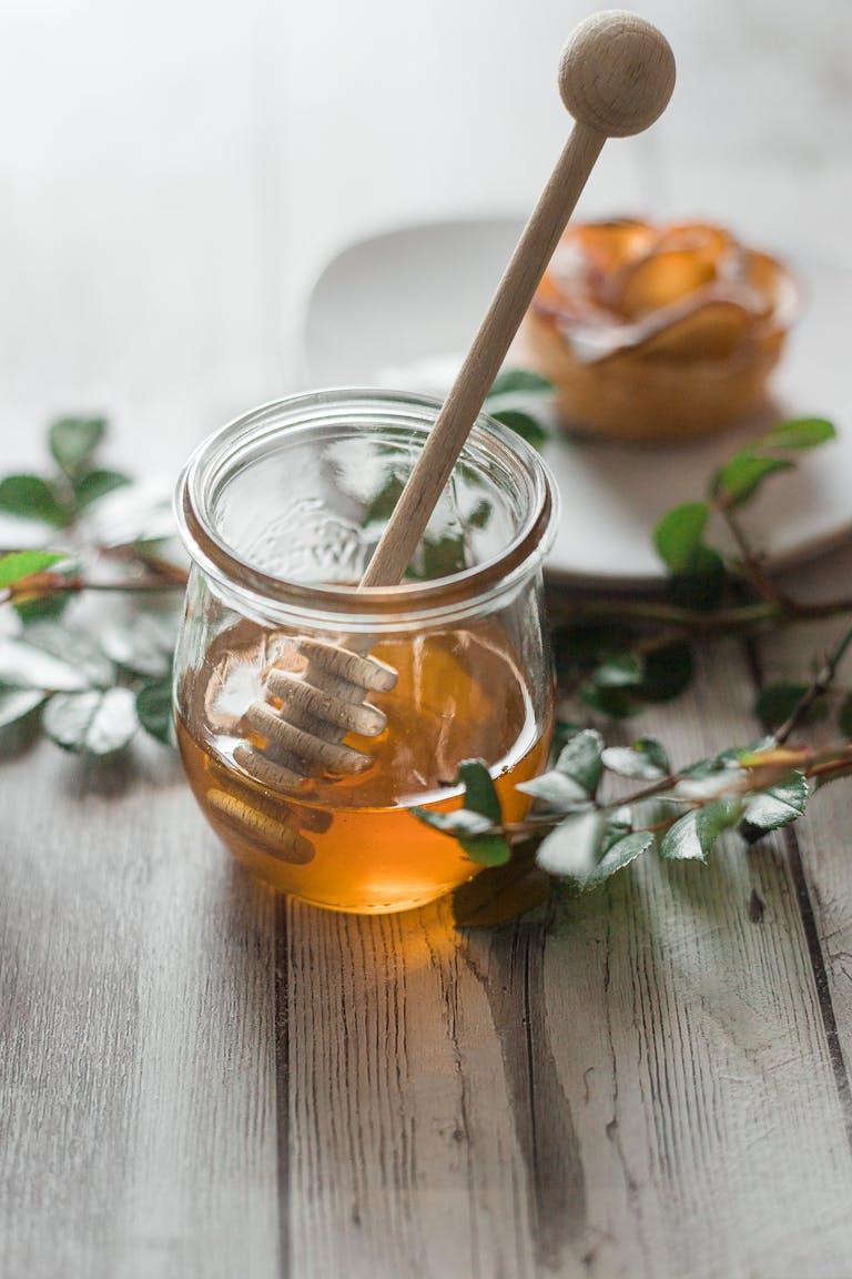 Glass jar filled with honey, featuring a wooden dipper, surrounded by greenery.