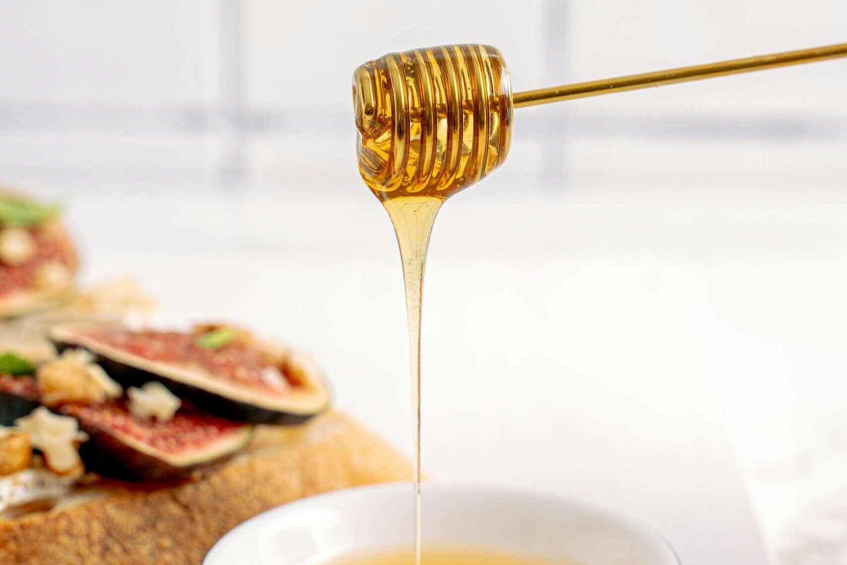 Honey flowing from dipper into bowl beside fig-topped bread on white table.